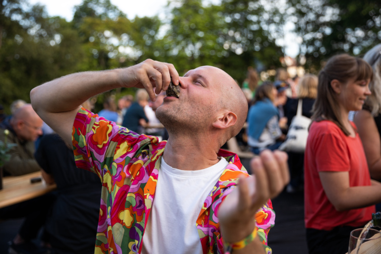 Bildet viser en skalla mann i fargerik hawaiiskjorte som bånner en østers. Han ser glad ut.